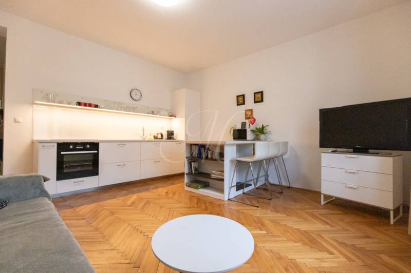 Nappali egy kétszobás lakásban konyhával, fa hatású padlóval, TV-vel és asztallal.