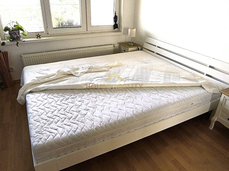 Világos hálószoba fehér ággyal és fa dekorációjú padlóval egy kétszobás lakásban.