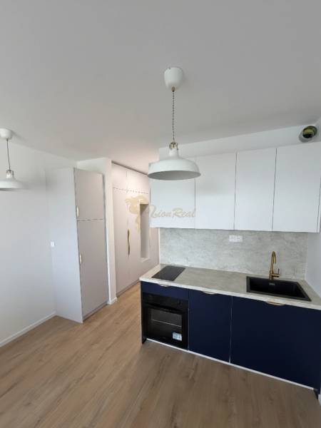 Egy garzonlakás konyhája világos szekrényekkel, szürke munkalappal és fa mintázatú padlóval.