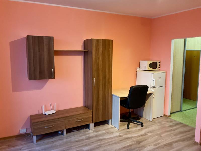 Garzonlakás bútorokkal, íróasztallal, mikrohullámú sütővel és fa mintázatú padlóval.