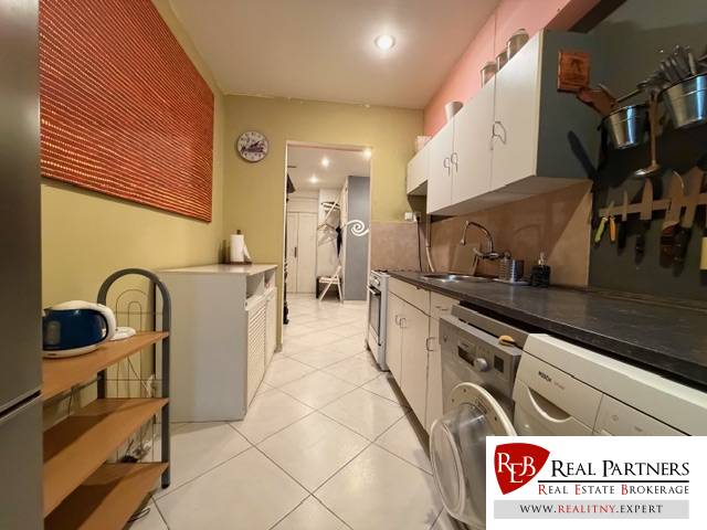 Ötszobás vagy annál nagyobb lakás konyhája fehér szekrényekkel és csempével, mindkét oldalon háztartási gépekkel.