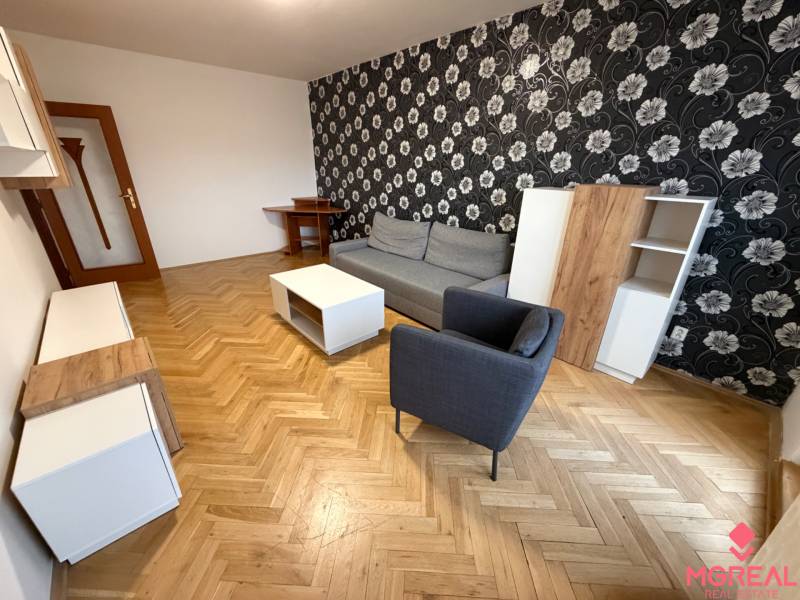 Nappali egy háromszobás lakásban fa mintázatú padlóval, mintás tapétával és bútorokkal.