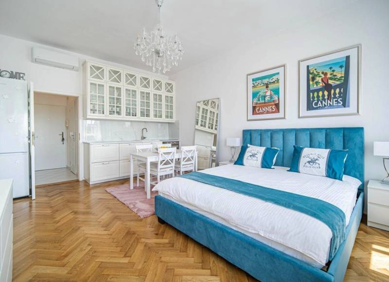 1 szobás lakás fa hatású padlóval, ággyal és fehér konyhaszekrényekkel.