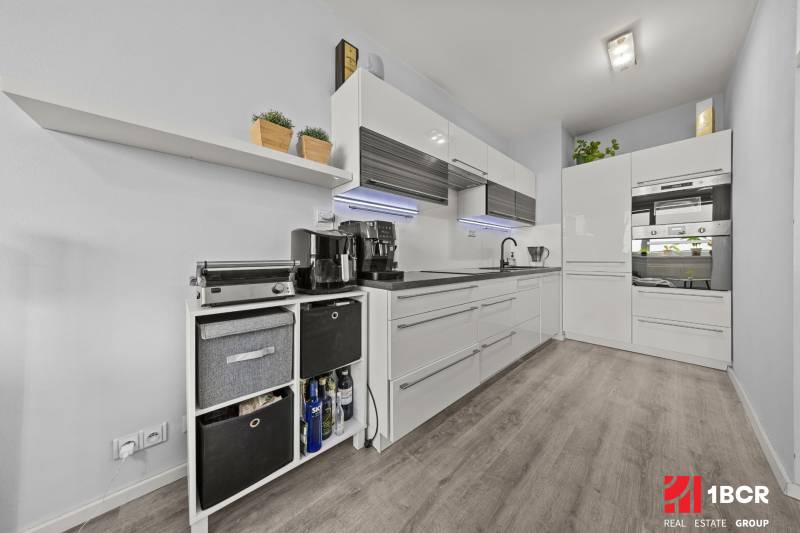 Konyha egy kétszobás lakásban fehér szekrényekkel és fa mintázatú padlóval.