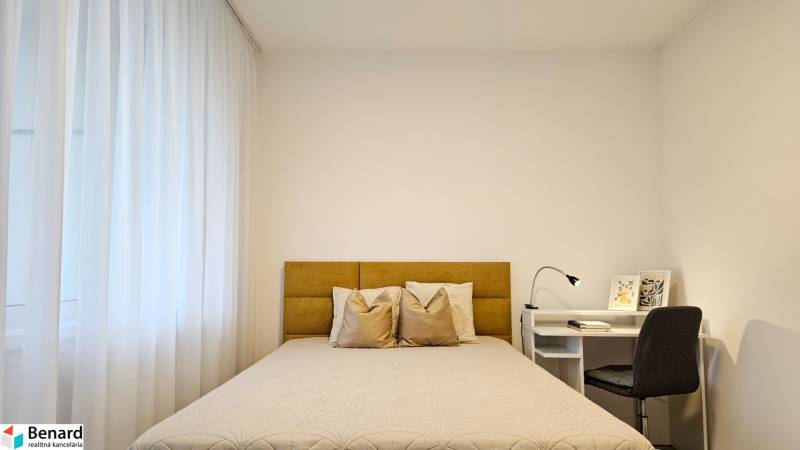 Hálószoba egy egyszobás lakásban világos ággyal, függönyökkel és fehér asztallal, lámpával.