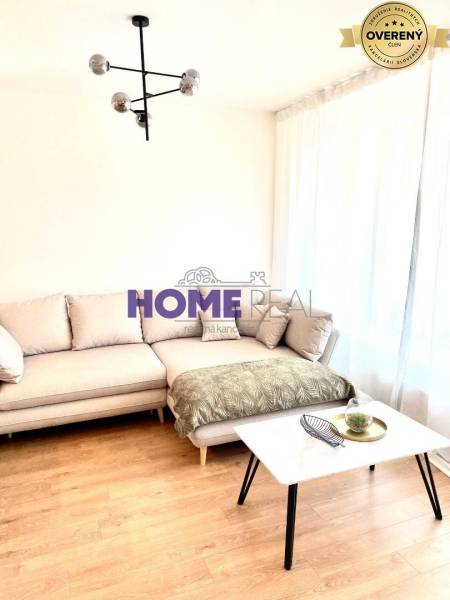 Nappali egy háromszobás lakásban kanapéval, asztallal és fa mintázatú padlóval.