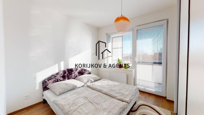 Világos hálószoba nagy ággyal, ablakkal és fa dekorációjú padlóval egy kétszobás lakásban.