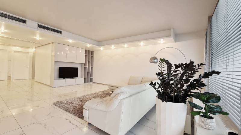 Nappali egy háromszobás lakásban fehér kanapéval, növényekkel és televízióval a világos padlón.