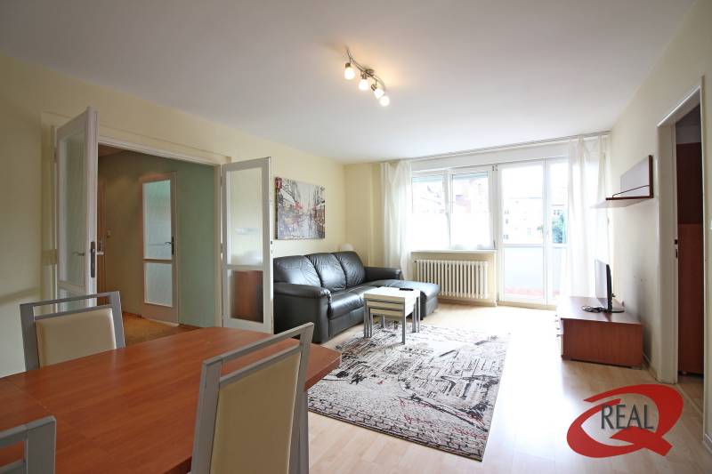 Nappali egy háromszobás lakásban fa mintázatú padlóval és fekete kanapéval.