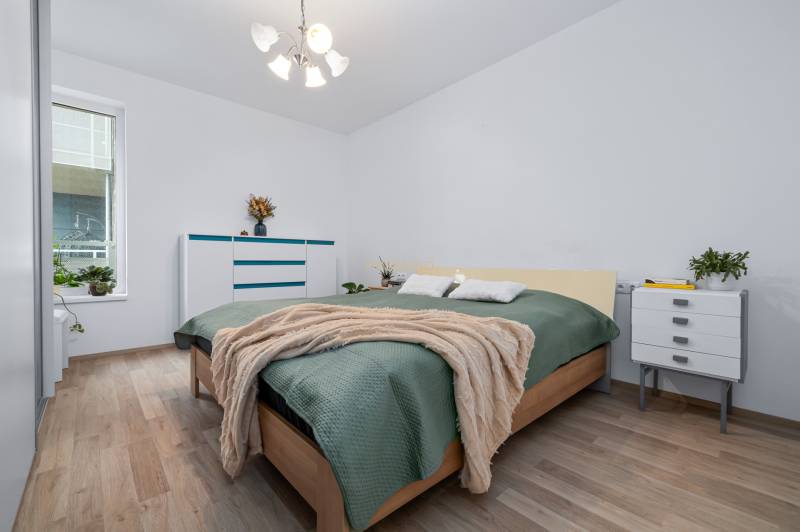 Hálószoba ággyal, komóddal, növényekkel és fa dekorációjú padlóval egy háromszobás lakásban.