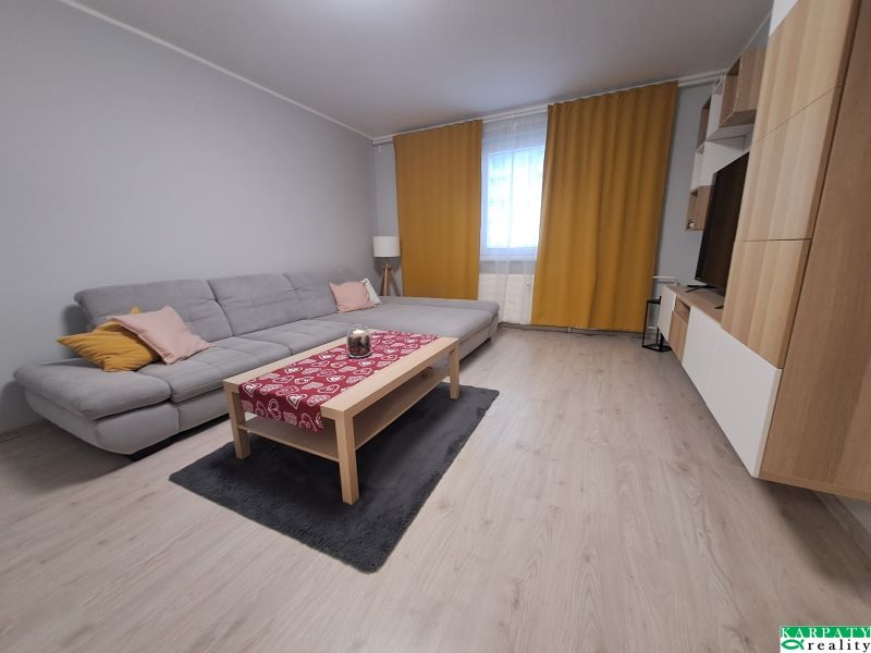 Nappali egy háromszobás lakásban kanapéval, függönyökkel és fa mintázatú padlóval.