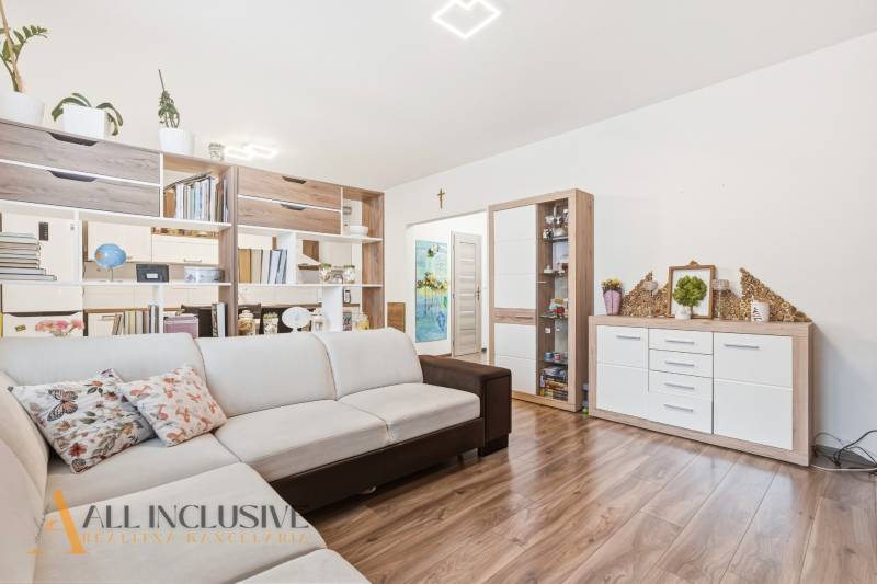 Nappali kétszobás lakásban bézs kanapéval és fa mintázatú padlóval.