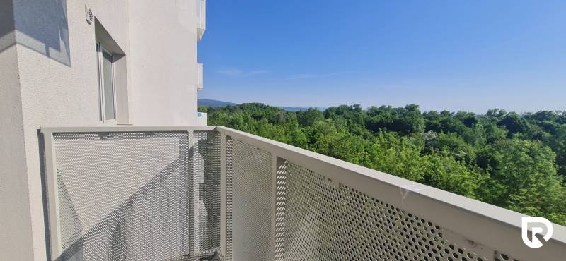 Erkély kilátással az erdőre és dombokra, háttérben egy épület, tiszta égbolt.