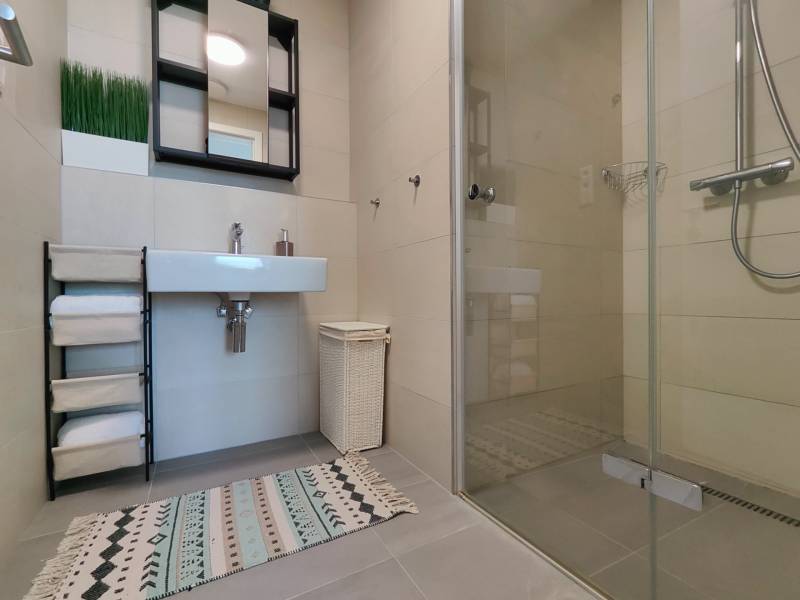 Fürdőszoba egy kétszobás lakásban mosdóval, tükörrel, zuhanyzóval és szőnyeggel.