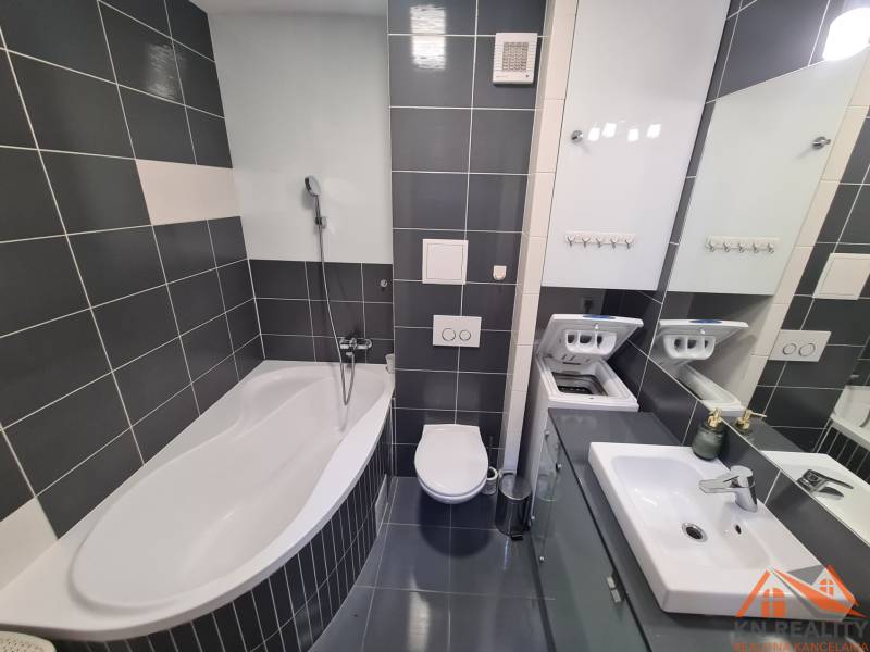 Fürdőszoba egy kétszobás lakásban káddal, mosógéppel, mosdóval és sötét csempékkel.