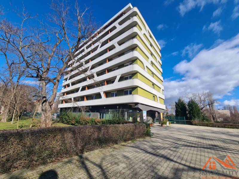 Egy hét emeletes lakóház Komáromban, fákkal körülvéve és burkolt járdával.