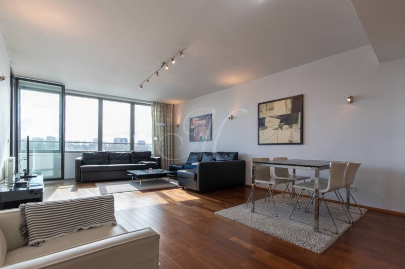 Nappali nagy ablakokkal, fa dekorációjú padlóval, kényelmes bútorokkal és étkező sarokkal.