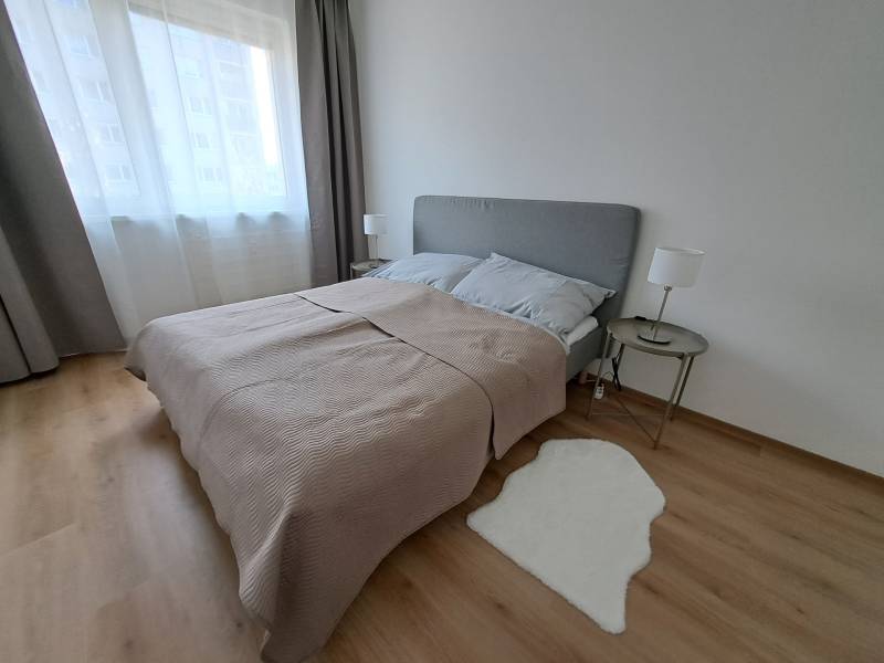 Hálószoba egy háromszobás lakásban, fa dekoros padlóval, szürke ágytámlával és szőnyeggel.