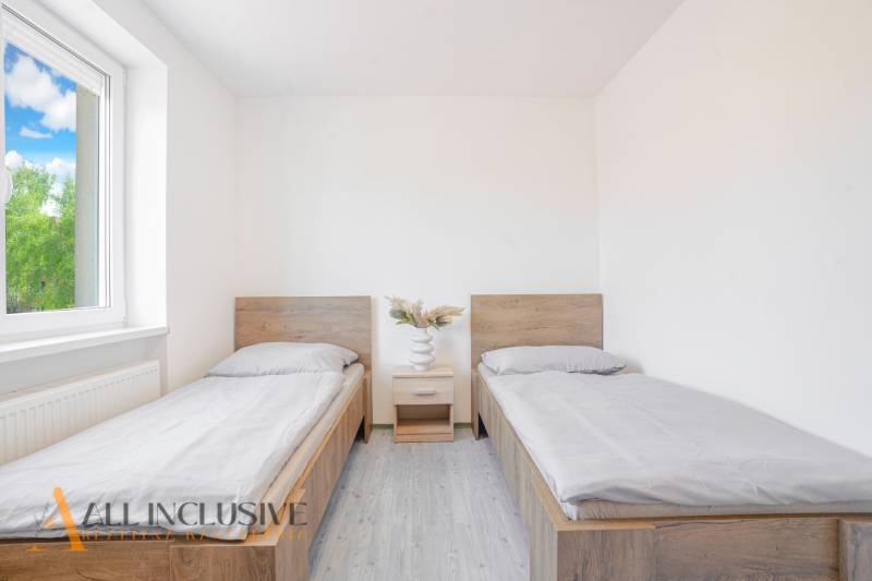 Szoba két ággyal, éjjeliszekrénnyel és fa mintázatú padlóval.