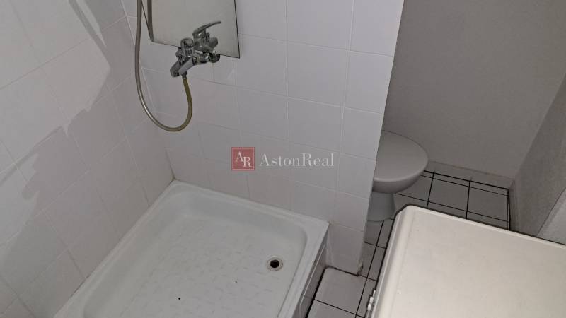 Fürdőszoba egy egyszobás lakásban fehér csempével és zuhanyzóval.