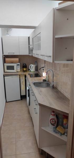 Keskeny konyha egy egyszobás lakásban, konyhapulttal, készülékekkel és tárolószekrényekkel.