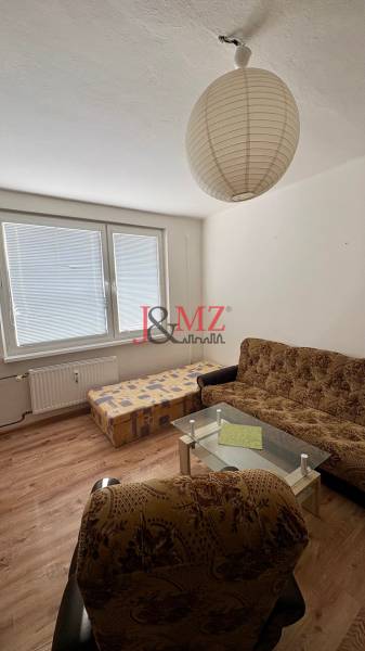 A garzonlakás tartalmaz egy kanapét, egy fotelt, egy dohányzóasztalt, fa dekorációjú padlót és egy nagy ablakot.