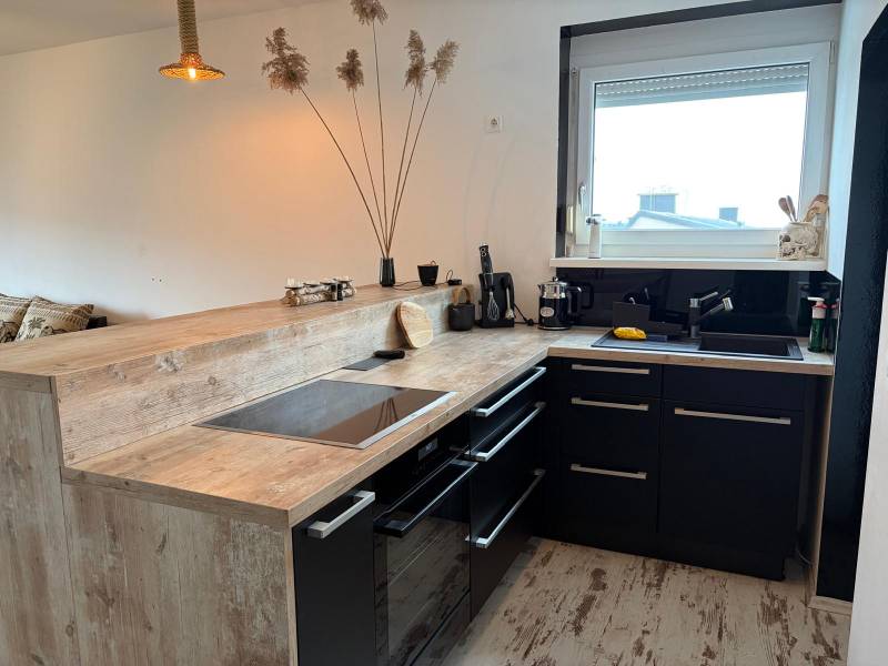 Konyha egy háromszobás lakásban fekete szekrényekkel és fa mintázatú padlóval.