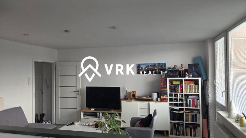 Nappali egy háromszobás lakásban könyvespolccal, televízióval és világos bútorokkal.