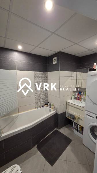 Fürdőszoba egy háromszobás lakásban káddal, mosógéppel és sötét csempékkel.