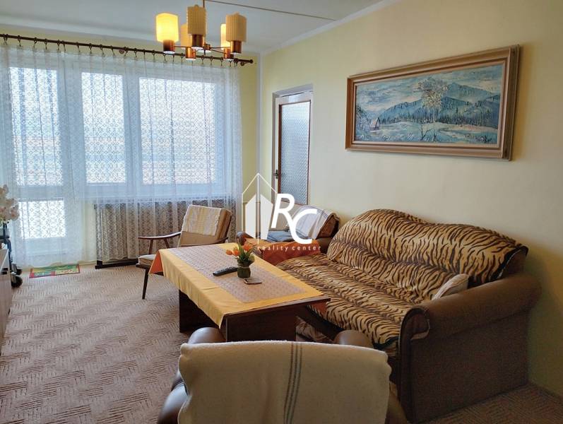 Nappali kanapéval, fotellel és asztallal egy kétszobás lakásban nagy ablakkal.