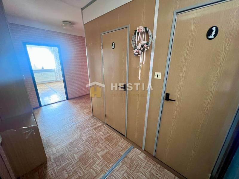 Folyosó egy 3 szobás lakásban, fa mintázatú padlóval és díszes koszorúval az ajtón.