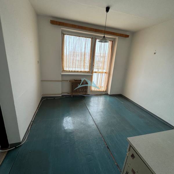 Egyszobás lakás egyszerű szobája ablakkal és radiátorral, sötét dekorú padlóval.