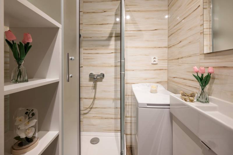 Fürdőszoba virágokkal, zuhanyzóval, mosdóval és tükörrel egy kétszobás lakásban.