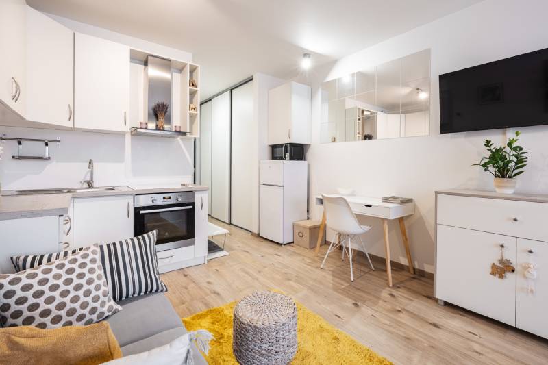 Nappali egy egyszobás lakásban konyhabútorral, fehér bútorokkal és fa dekorációjú padlóval.