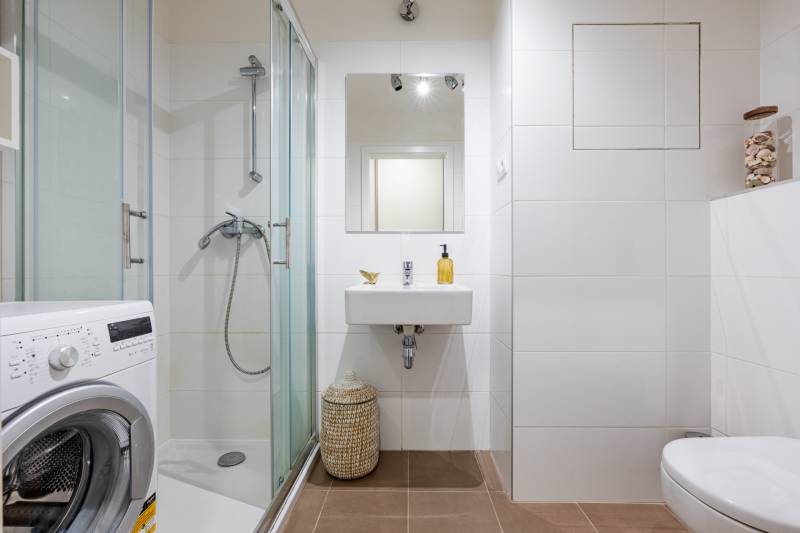 Fürdőszoba egy egyszobás lakásban mosógéppel, zuhanyzóval és fehér csempékkel.