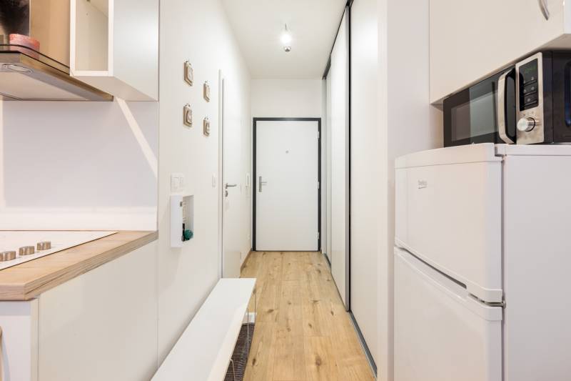 Keskeny folyosó konyhapulttal, mikrohullámú sütővel és fa dekorációjú padlóval egy egyszobás lakásban.