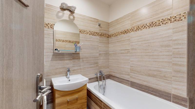 Fürdőszoba egy kétszobás lakásban világos csempékkel és káddal, mosdó alatti szekrényekkel.