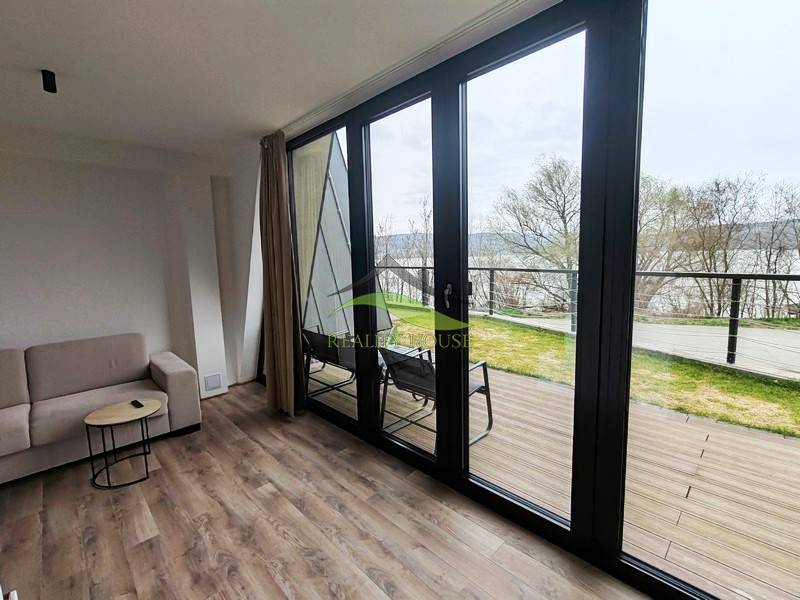 Ház belső tere kanapéval, fa dekorációjú padlóval, nagy ablakokkal, kilátással a teraszra.