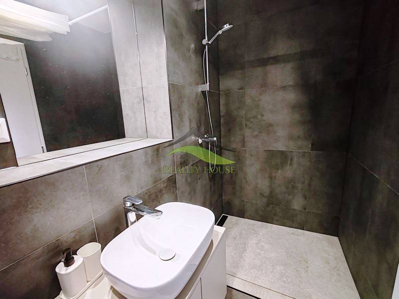 Fürdőszoba a házikóban mosdóval, tükörrel és zuhanyzóval, sötét csempékkel.