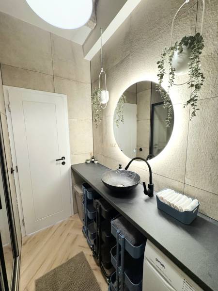 Fürdőszoba ívelt tükörrel, függő virágokkal és fa mintázatú padlóval.