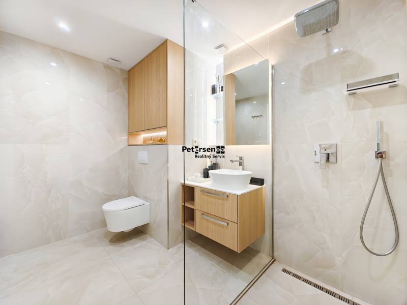 Fürdőszoba egy kétszobás lakásban, walk-in zuhanyzóval, fehér mosdóval és fa szekrényekkel.