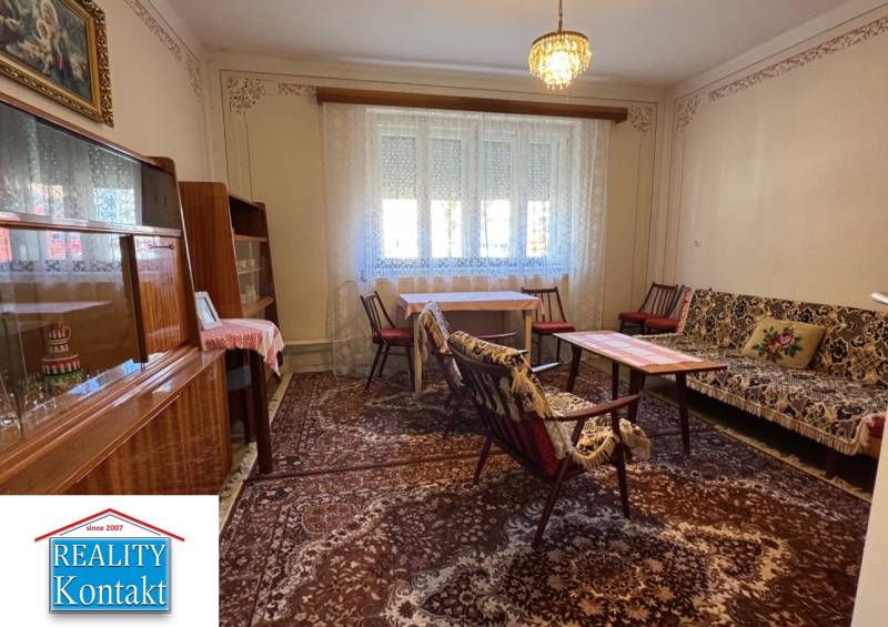 Nappali egy családi házban szőnyeggel, bútorokkal és csillárral, függönyök az ablakokon.