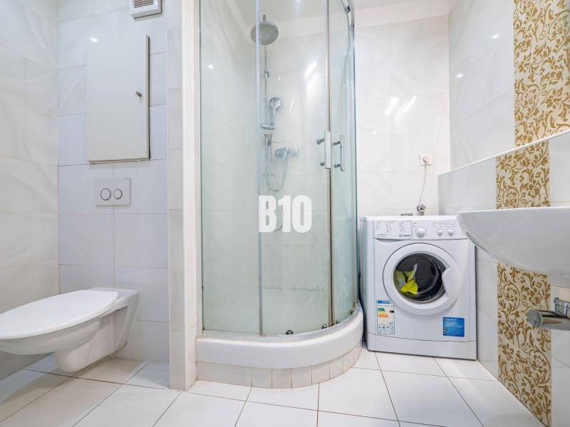 Fürdőszoba egy egyszobás lakásban mosógéppel, zuhanyzóval és kerámia csempékkel.