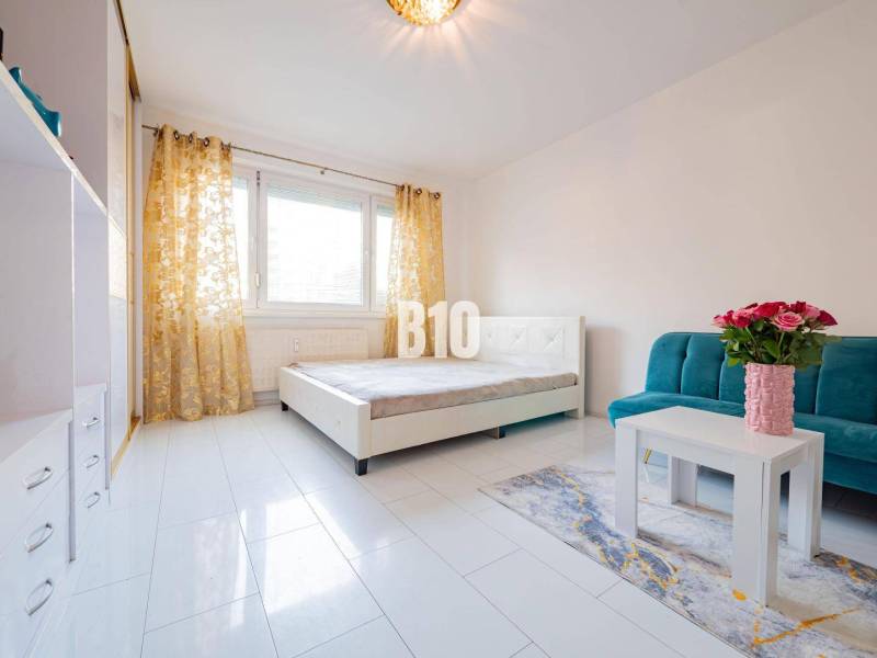 Világos szoba egy egyszobás lakásban kanapéval, asztallal és vázában lévő virágokkal.