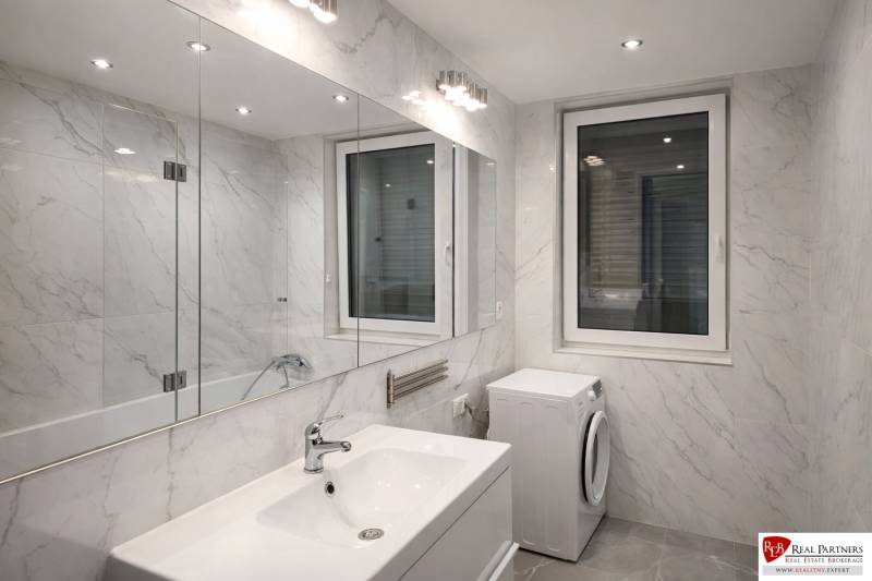 Fürdőszoba egy kétszobás lakásban káddal, mosdóval és mosógéppel, márvány kinézetű burkolattal.