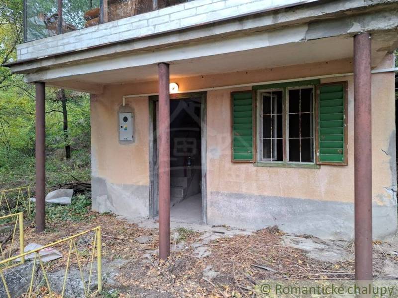 Kőház verandával és zöld zsalugáterekkel, természet által körülvéve.