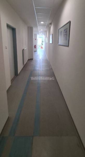 Hosszú folyosó fehér falakkal, mennyezeti lámpákkal és irodai képekkel.