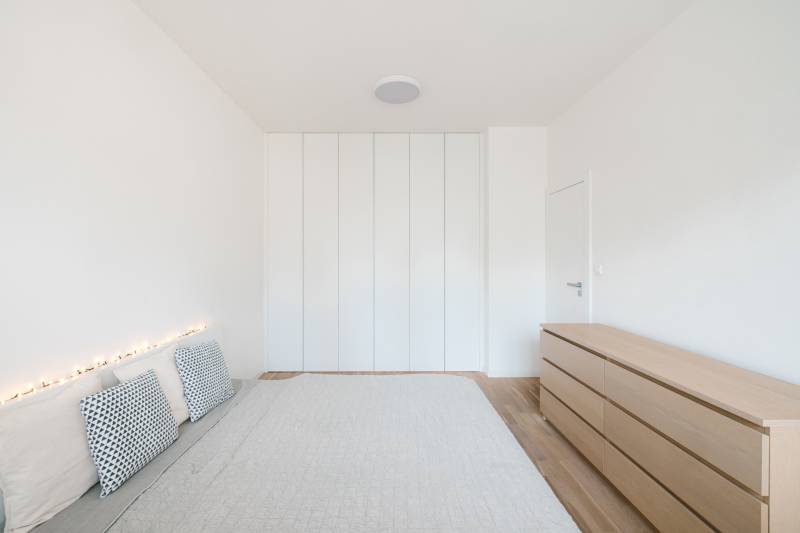 Világos hálószoba egy négy szobás lakásban, ággyal, párnákkal és szekrénnyel, fa dekoros padlóval.