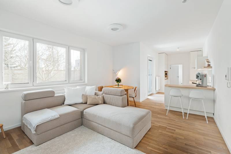 Világos nappali konyhasarokkal, kanapéval és fa mintázatú padlóval.