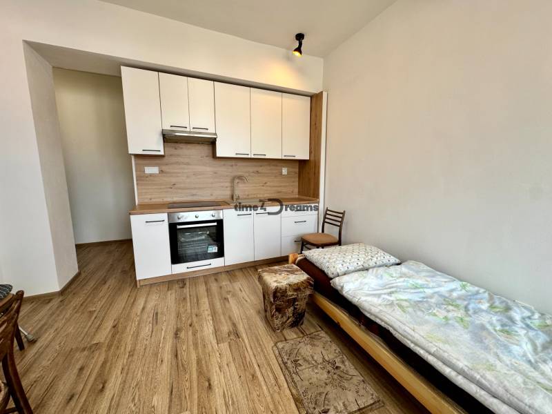 Garzonlakás konyhasarokkal és fa mintázatú padlóval, kiegészítve ággyal és székkel.